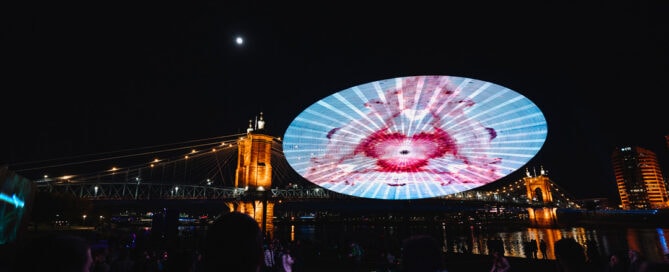 Vibrant nighttime scene of Cincinnati's Roebling Bridge illuminated with colorful digital art projection during BLINK, attracting a large crowd along the riverfront.