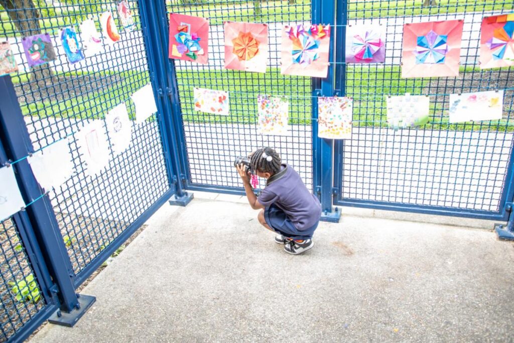 Child crouching and viewing colorful paper art pieces displayed on a blue metal fence at an outdoor arts event.