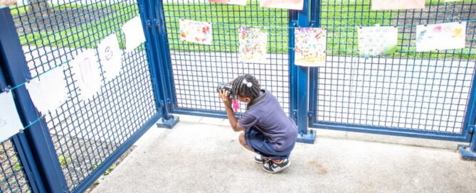 Child crouching and viewing colorful paper art pieces displayed on a blue metal fence at an outdoor arts event.