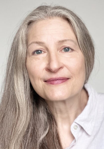 Close-up of a mature woman with long gray hair and blue eyes, smiling gently, wearing a white shirt, representing community and arts engagement.